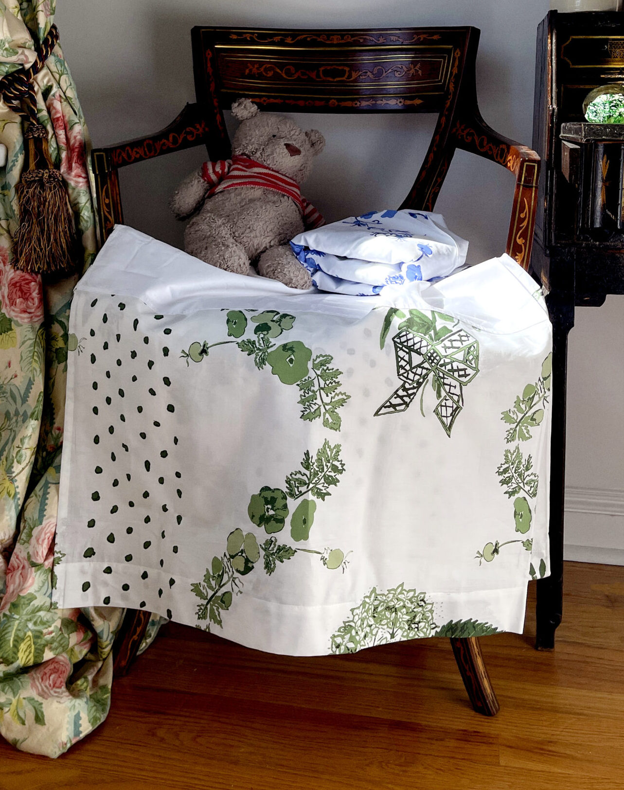 Two folded sheets on an antique chair with a teddy bear and a chintz curtain