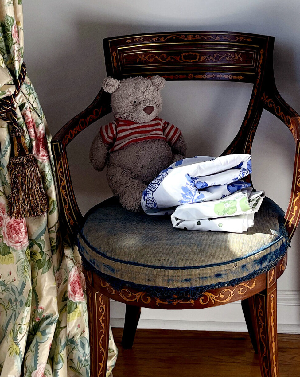 Two folded sheets on an antique chair with a teddy bear and a chintz curtain